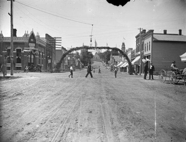 View down center of unpaved street towards men passing welcome arch for the Tenth Wisconsin in the middle of Main Street, between Water and First Streets. Jackson County Bank is on the left. The flood of 1911 cut off the bank, except the vault. The bank was moved next door and an opening cut into the vault. On the right is the Jones and Murray Hardware Store. Next was Eckern's Jewelry. Eckern was a hunchback and bachelor, well-known for his kindness and generosity. The store also handled schoolbooks and stationery.