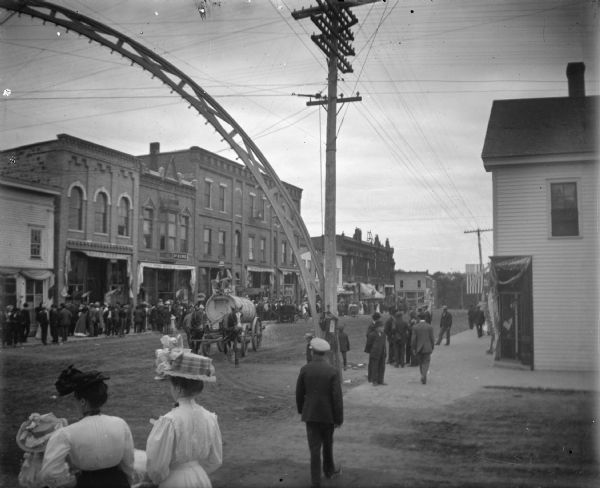 Slightly elevated view towards a horse-drawn water sprinkler on a wagon approaching an arch over the intersection during a parade. People are gathered on the sidewalks.