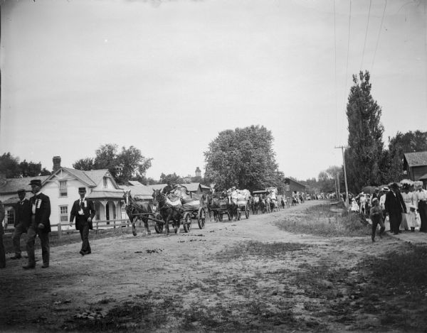 View down side of street towards a parade of men and women in patriotically decorated wagons coming up a road outside of town. People are watching from the boardwalk on the right.