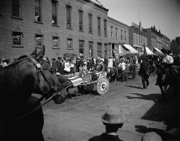 Slightly elevated view of a parade of men and women in patriotically decorated wagons going through town. A horse is in the foreground on the left, and a man wearing a hat is in the center foreground.