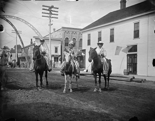Three patriotically dressed women on horseback pose on an unpaved street in town. The large flags have 45 stars, and the small flags have 46 stars. In the background is an arch over the intersection.