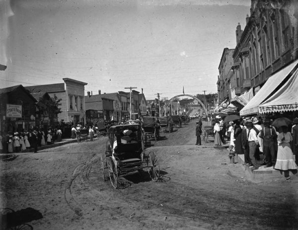 Slightly elevated view of buggies following marchers in parade though town, making a right turn from Water Street to Main Street and heading west. In the background is an arch over the intersection. A large poster reads" "Wisconsin State Fair, 1908, September 7, 8, 9, 10, 11."