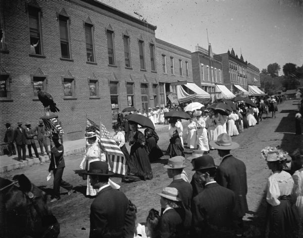 Women's Relief Corps with umbrellas parading down the street. Stuffed eagle parading as "Old Abe."  Directly behind the two flags are, from left to right: Grandma Maddox and Mrs. Frank Cooper, holding umbrella and looking at the camera. Memorial Day parade.