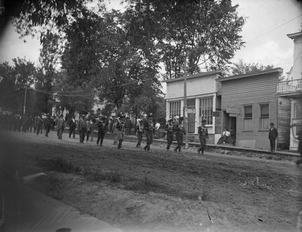 Men parading down street with axes over their shoulders. Probably the Modern Woodmen of America, a fraternal insurance body in the 1890s which was strong in number; its rates were very low.