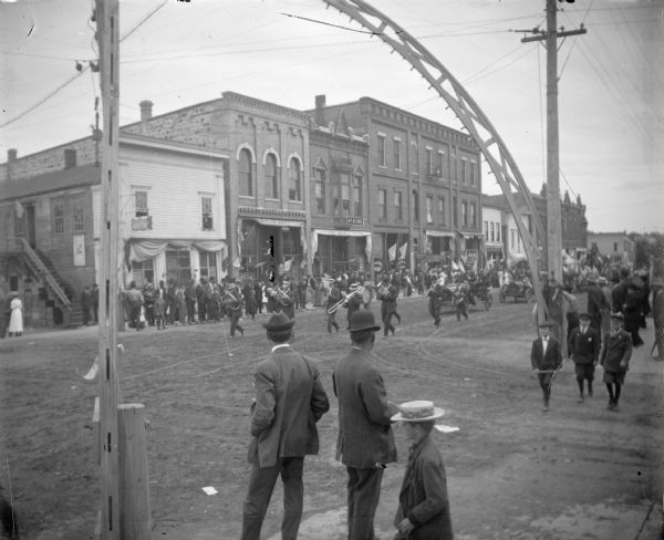 View from sidewalk towards a parade with band and automobiles going up hill on Main Street past the arch over the intersection.