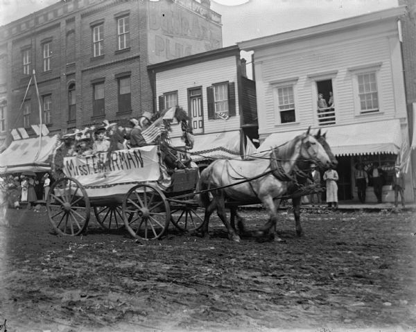 Horse-drawn wagon carrying people wearing costumes and hats in a parade. The banner on the side reads, "Miss F.E. Harman, who ran a milliner's establishment.