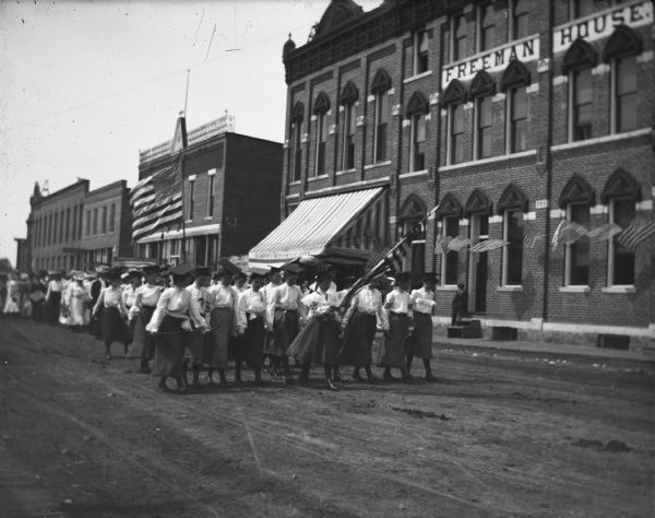 Parade of women passing the Freeman House, wearing mortarboards and carrying a Union High School flag with the date 1897.