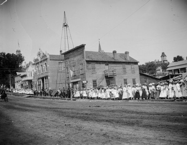 Parade of women and children on sidewalk through town. They are going past Peter Olson's blacksmith shop (foreground), fire-bell tower, and the City Hall with library upstairs. This is probably a procession to the cemetery on Memorial Day, with people carrying wreaths. The occasion was locally called "Decoration Day". It was a mile-and-a quarter walk to the cemetery.