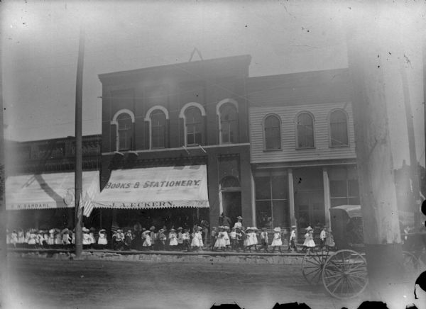 Parade of children in procession on the board sidewalk in front of J.A. Eckern's store, probably Memorial Day.