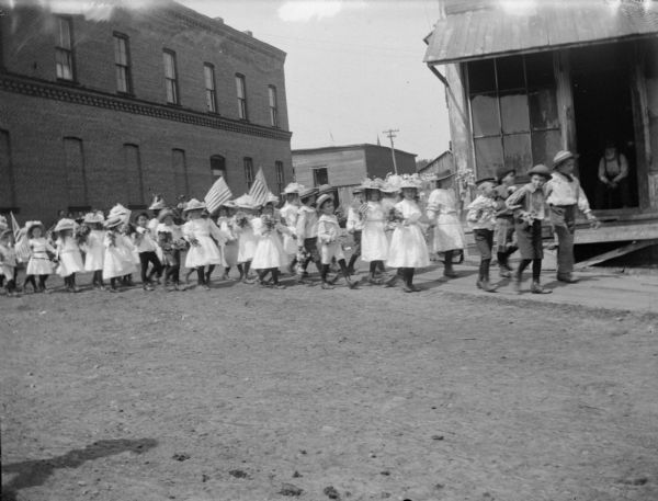 Small children parade with flags and flowers in a Memorial Day procession. The children were kept in formation by each child holding the end of a wooden crosspiece tied to a line of rope.