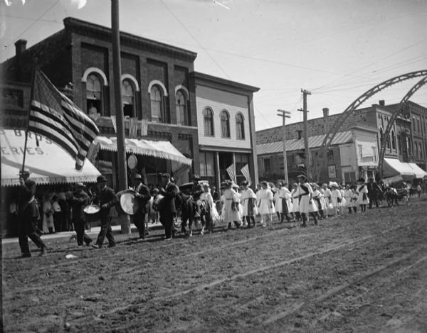 Band following a flag with Union High School embroidered on it, parading west on Main Street, followed by a group of women. In the background on the right is an arch over the intersection