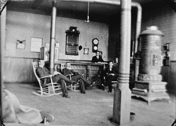 Interior view of men sitting in rocking chairs around a wood stove, probably inside a hotel/inn.