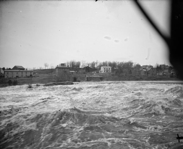 View of the river and town. High water looking west. Site of the present dam, but this was the old roller dam. Building with an advertisement for Veterinary Carbolisalve for Horse & Cattle right of center.