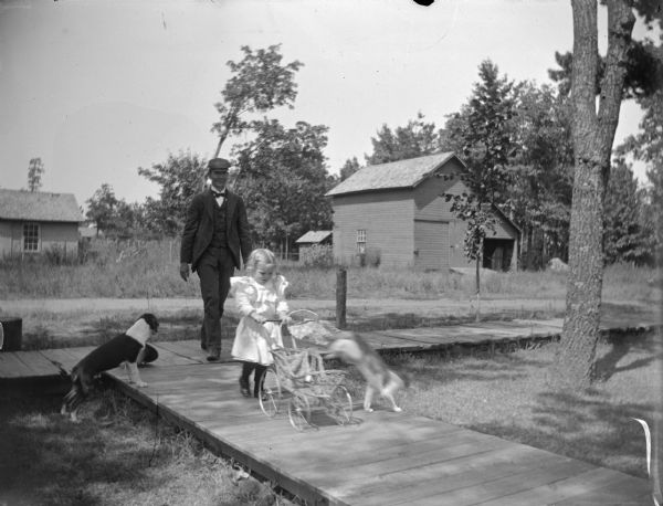 Cat jumping out of a baby carriage pushed by a girl on a board sidewalk. She is followed by a man and dog. Probably Rufus A. Jones and daughter.