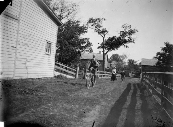 Man riding a bicycle on a residential street, carrying packages in his left arm. Behind him is a woman and a girl walking behind. Fences are along the side of the street, and barns are on the left and in the background.
