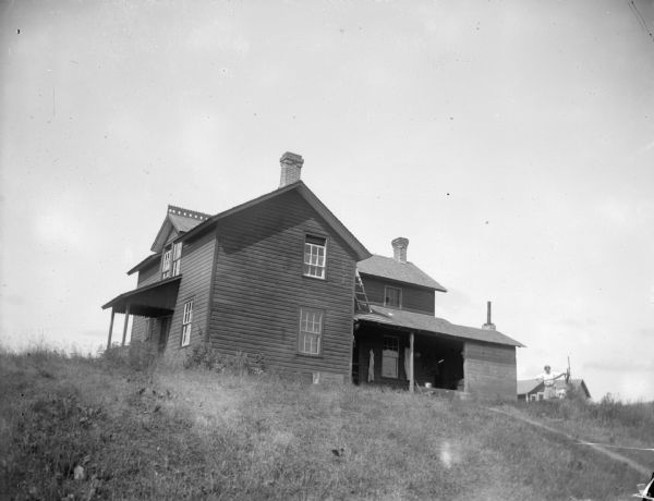 View up hill towards farmhouse. There is a woman using a hand-pump in the yard on the right.