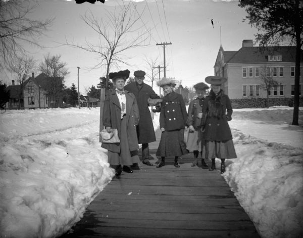 Group of four women and man posing on a board sidewalk between snow drifts in front of the high school.