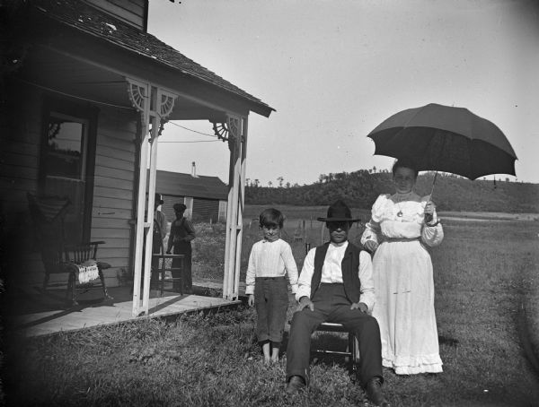 Seated man, probably William O'Brien, a woman with umbrella, Maggie Beatty O'Brien, and William Miles O'Brien, posing in front of a house. Two men are in the left background.