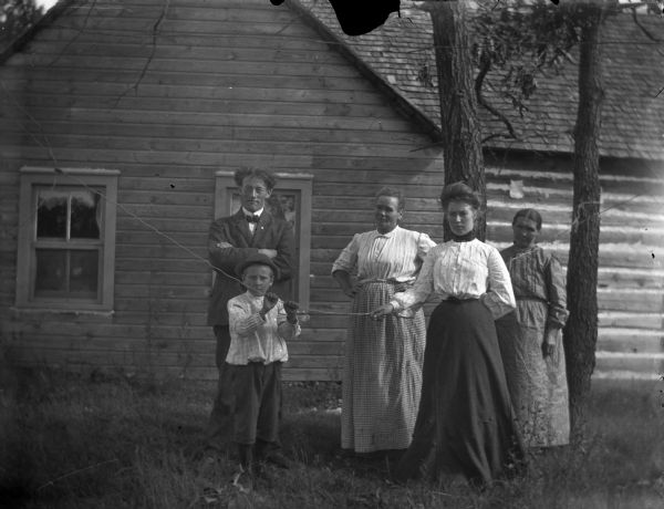 Five people, possibly family, posing in front of a building. A man is standing on the left with his arms crossed, and two women stand on the right near trees. A woman and a young boy in the foreground are holding onto a wire strung across the yard. 