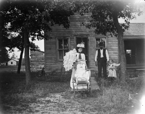 Man and woman, with umbrella, posing with a girl and baby in a wicker baby carriage in front of a frame house, number 419.
