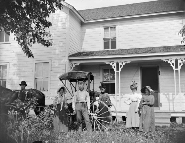 Group of people in front of a frame house with a top-buggy. Four women and two men The man on the far left is holding a bicycle.
