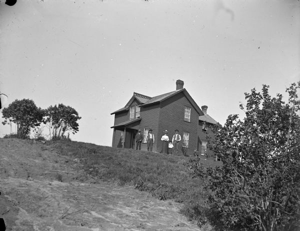 Group of three men, woman, and a boy posed in front of house on hill.
