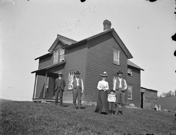 Group of three men, woman, and a boy posed in front of house on hill.