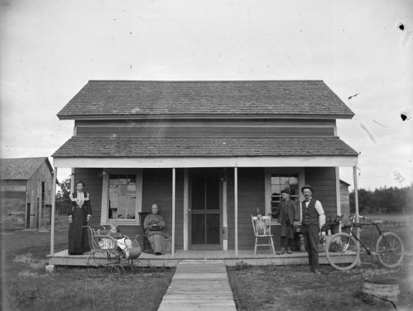 Group of people posed in front of a frame house. Two women, one seated, and a child in a buggy on the left side; two men, a seated dog, and a bicycle on the right.