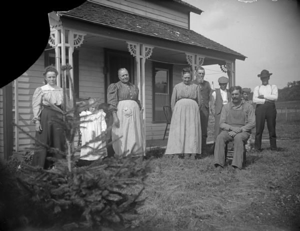 Three women, four men (one seated), a boy, and a girl posing in front of a house.