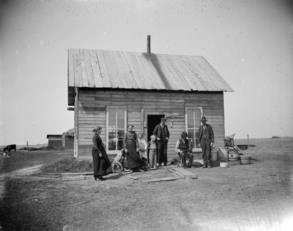 Group of people and a dog in front of small frame house. One of the men is holding a violin.