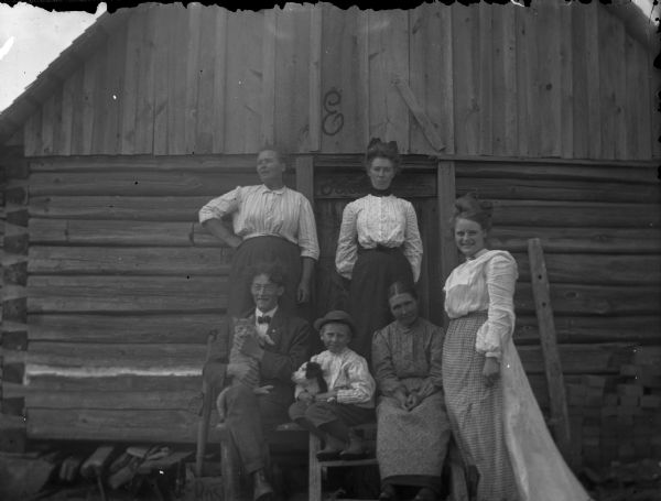 Group of four women, man, and boy in front of a log building. The man is holding a cat, and the boy is holding a dog.