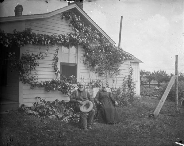 Man and woman sitting in front of a morning glory vine-covered frame house, possibly Irving Nutter and his wife.