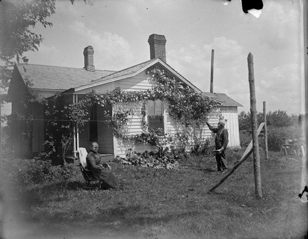 Man posed standing and a seated woman in front of a morning glory vine-covered frame house, possibly Irving Nutter and his wife.
