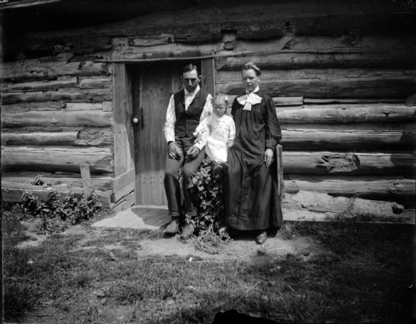 Man, woman, and girl posed in front of a log cabin.
