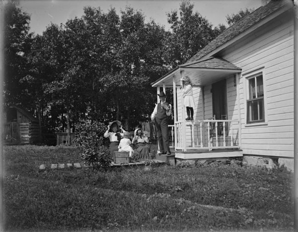 A man playing a violin is standing near a woman and three children in front of a frame house.
