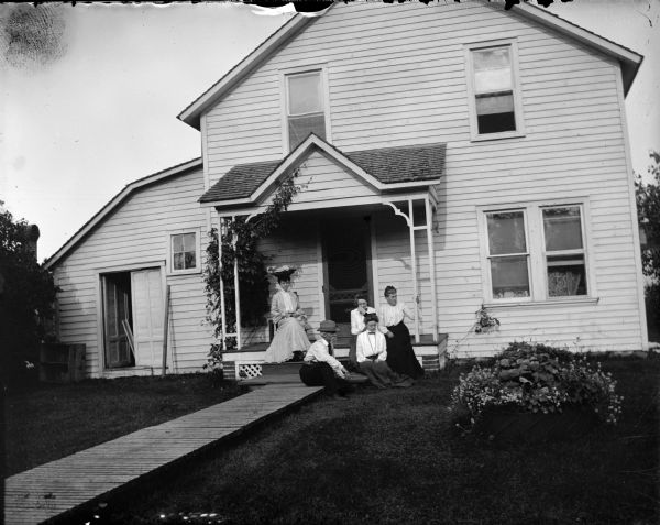 Four women and a man seated on the front porch of a frame house.