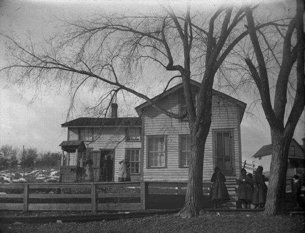 Group of girls passing by a frame house with a well in the yard. Two women and a man pose in the left doorway.
