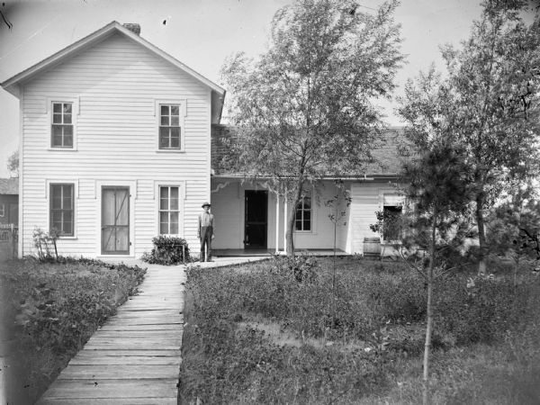 Man posing on the board sidewalk in front of a frame house.