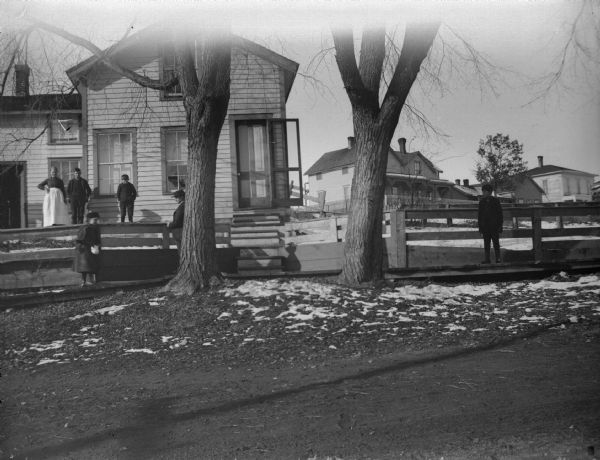 Man, woman, and boy posing in front of a frame house, while a man, girl, and boy pass on the board sidewalk.
