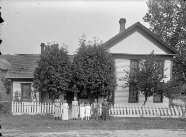 Four women, a man, and two girls posing in front of a two-story and one-story house.