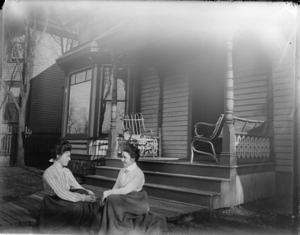 Two women sitting on the board sidewalk in front of a house.