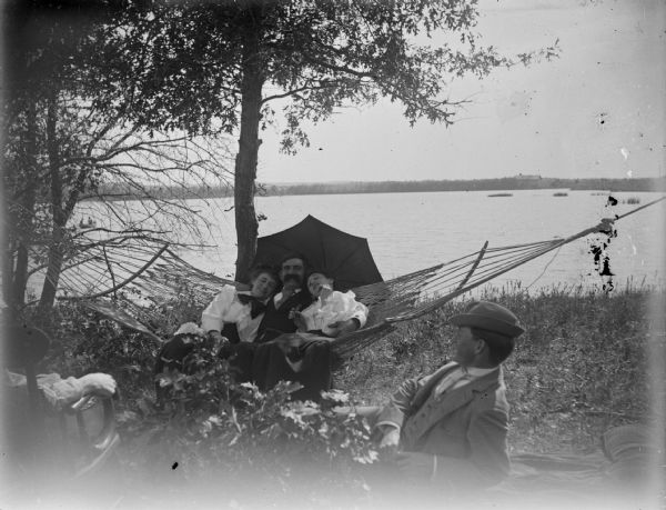 Man and two women in a hammock down by the river. Another man reclining on a blanket.