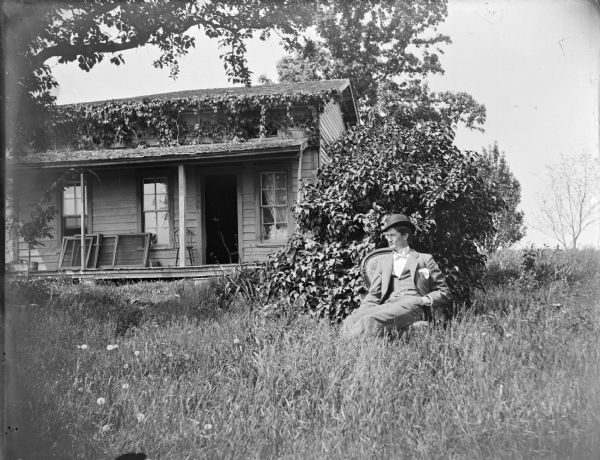 Outdoor view of a man wearing a hat sitting in the yard in front of a frame house.