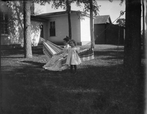 Woman sitting in a hammock and a girl standing in a backyard.