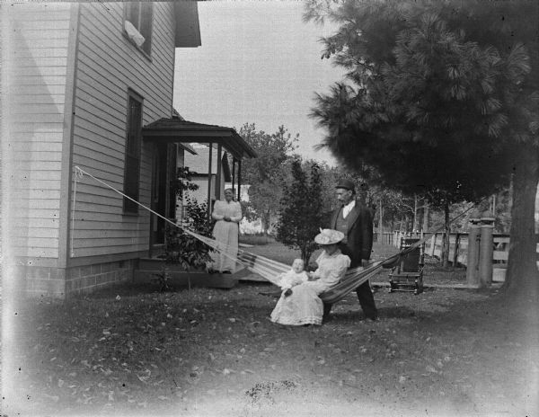 Woman and child sitting in a hammock. A woman and man stand behind them in the yard of a house.