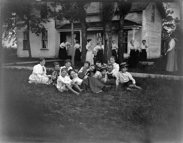 Women standing on porch and near a house among trees, with children in the foreground sitting on the lawn. One woman is pulling a wagon. R. Rainey home, S.E. corner of 6th and Pierce. From left seated: Cleo Preston, Oce Thompson; lying in front: Alice Robie; girl with hat: Grace Jones Mick; behind her: A. or M. Murray; standing in white shirt: Spaulding twin; standing 2nd from right: Grace Ogden.