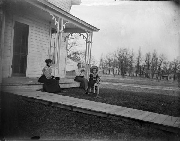 Two women are sitting on a porch, one holding a baby in her lap. A young girl wearing a hat is riding a rocking horse in the yard. A board sidewalk leads up to the porch.