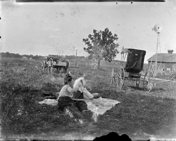 Two women playing with a baby while reclining on a blanket in a farm field with two carriages in the background.