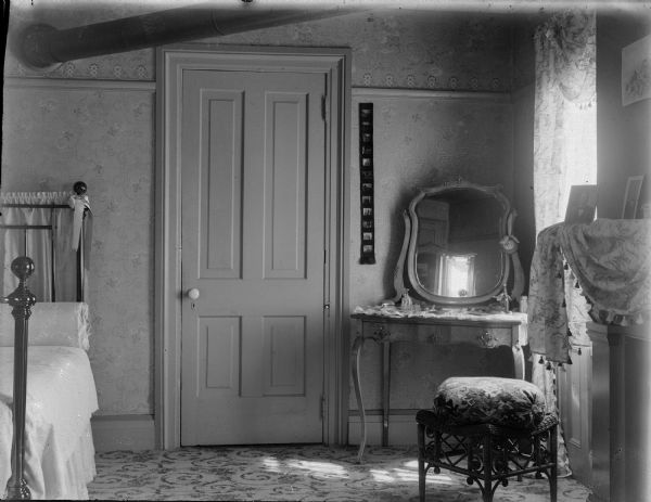 Interior of bedroom with bed, door, dressing table, and mantle.
