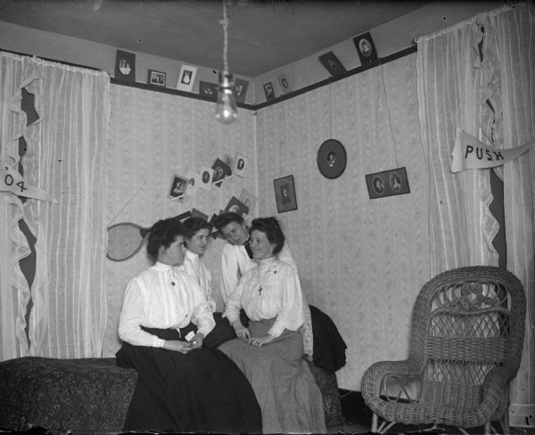 Four women sitting on a bed, walls displaying a tennis racquet and photographs.